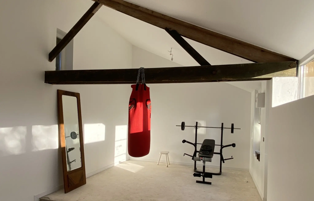 Salle de sport sous charpente avec banc, haltères et sac de frappe, au gîte Le Clos de l'église